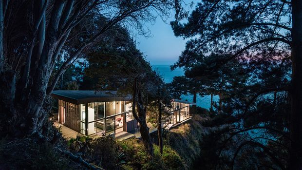 A compact, modern glass-walled pavilion or house nestled among dense coastal trees at dusk. Warm interior lighting reveals an open living space, while a narrow glazed extension or walkway projects toward a visible blue ocean or water view. The building has a low, flat roof and dark slender structural framing, blending into the wooded landscape.