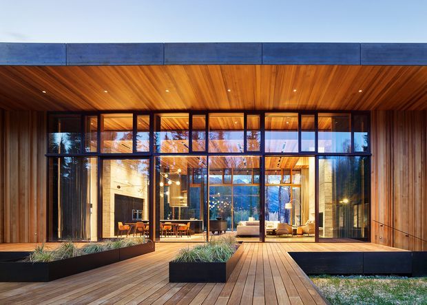Contemporary single-storey residential courtyard or entry facade with extensive black-framed glazing, warm timber soffits and wall cladding, and a timber deck walkway leading into the home. The glazed interior reveals a living and dining area, while integrated planters and dark raised garden beds define the approach.