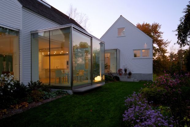 Exterior view of a light-coloured weatherboard house at dusk, featuring a prominent glazed sunroom or contemporary glass extension connecting two pitched-roof volumes. Warm interior lighting reveals a kitchen and dining area through the floor-to-ceiling glazing. The house is surrounded by a landscaped garden with lawn and flowering planting.