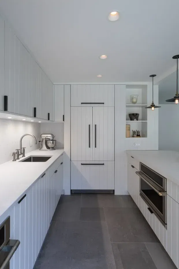 A bright, compact contemporary kitchen with white Shaker-style cabinetry featuring vertical groove detailing, long black bar handles, white benchtops, a stainless-steel sink and tapware, integrated oven, and open display shelving. Dark grey large-format floor tiles provide contrast, while recessed ceiling lights and black pendant lights illuminate the space.