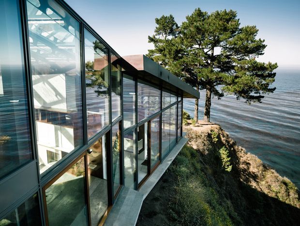 Exterior architectural view of a contemporary glass-walled residence projecting along a steep waterfront cliff. Extensive floor-to-ceiling glazing reveals a bright multi-level interior, while dark metal framing and a low, angular roofline contrast with the rugged coastal landscape and ocean beyond.