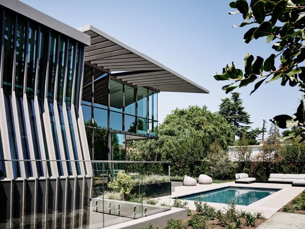 Contemporary multi-level residence with extensive floor-to-ceiling glazing, a sharply projecting slatted roof canopy, and a distinctive vertical fin facade. The house opens onto a landscaped outdoor area with a rectangular swimming pool, paved terrace, glass balustrades, and minimalist outdoor seating. Bright, refined architectural photography with strong indoor-outdoor connection.