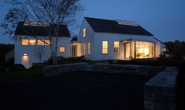 Twilight exterior view of a substantial white-sided house with multiple pitched gabled roof forms, dark roofing, illuminated rectangular windows, and a curved glazed sunroom or projecting living space. Low dry-stone retaining walls and landscaped grounds frame the paved driveway and entry approach.