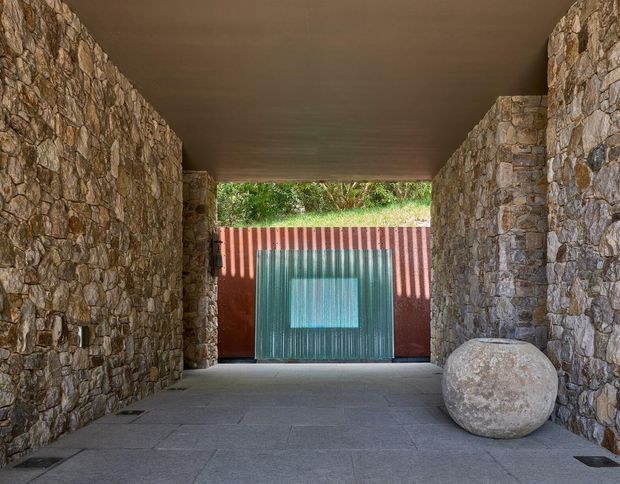 Covered entry passage framed by rough natural-stone walls, with a dark flat ceiling, large grey stone paving slabs, and a corrugated metal gate in muted red and teal at the far end. A large rounded stone is placed along the right wall, while greenery is visible above the gate.