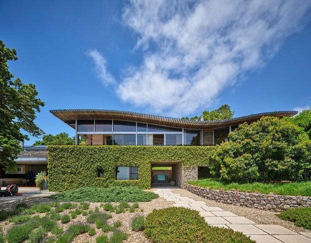 A contemporary two-level residential facade set within a landscaped garden. The lower level is largely concealed by a dense clipped hedge, with a central timber entry door and small windows. The upper level features extensive glazing, dark-framed windows, and a broad shallow projecting roof with closely spaced timber slats. Natural stone walls and dry-stone retaining walls integrate the house with the planted landscape. A paved path leads toward the entrance, while mature trees and a bright blue sky frame the scene.