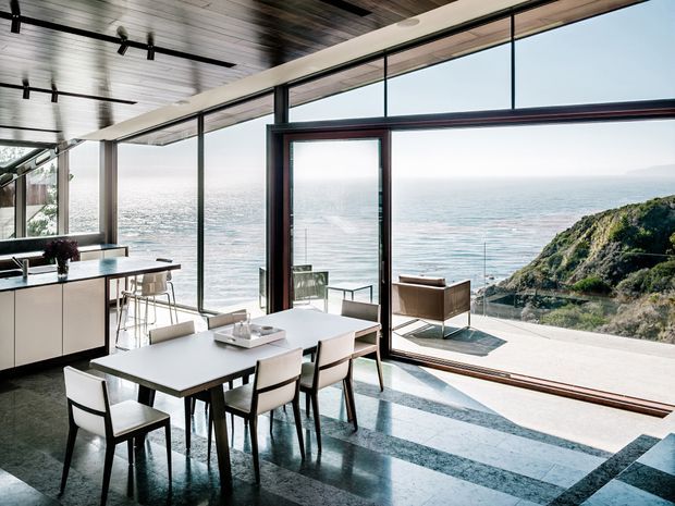 A bright, open-plan dining and kitchen area with full-height glazing overlooking the ocean and a rocky coastal headland. The room features a large rectangular dining table, upholstered dining chairs, minimalist kitchen cabinetry with a peninsula and stools, dark polished stone flooring with lighter geometric bands, exposed dark timber ceiling boards, and slim dark window and door frames. A large glazed door opens directly to an outdoor terrace.