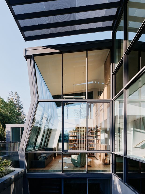 A dramatic view of a contemporary multi-level residential extension or courtyard facade, dominated by black-framed floor-to-ceiling glazing and a large translucent roof canopy. The glazed volume reveals a double-height interior with timber flooring, a sculptural lounge chair, shelving and a staircase, while angled glass walls and exposed steel framing create a highly geometric composition. Trees and sky are visible beyond the building.
