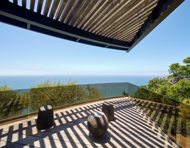 A sunlit elevated outdoor terrace overlooking the ocean, with a timber-slatted pergola casting strong linear shadows across the deck. The terrace is enclosed by frameless glass balustrades and contains two dark sculptural stools or side tables, with coastal vegetation at the edges.
