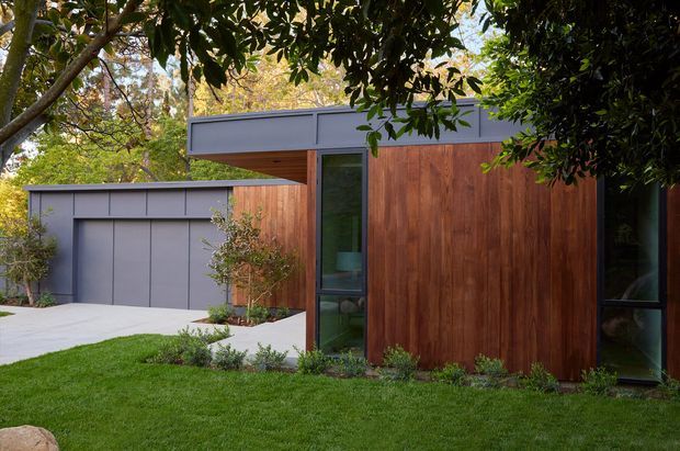 Exterior view of a contemporary single-storey residence or ancillary building, featuring warm vertical timber cladding, dark grey metal-clad flat roof forms, large slender glazed openings, and an attached matching garage. The building is set within a landscaped garden with lawn and mature trees.