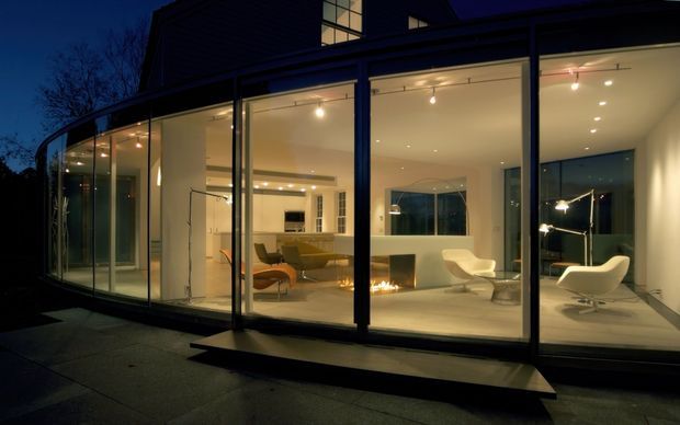 Night-time exterior view of a contemporary glazed house extension. Floor-to-ceiling glass walls reveal a warmly lit open-plan living area with modern lounge seating, a fireplace, and a kitchen beyond. Dark structural framing and a curved glazed frontage contrast with the illuminated minimalist interior.