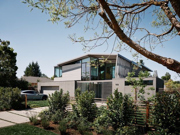 Exterior view of a contemporary two-storey residence with a low-pitched asymmetrical roof, extensive dark-framed glazing, horizontal metal or fibre-cement cladding, and a dark neutral ground floor. The house is screened by layered planting and a horizontal timber fence, with a concrete driveway and a carport or garage visible at left. The suburban setting and mature street tree frame the composition.
