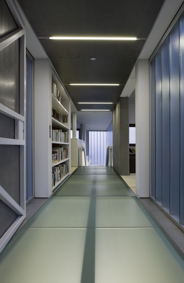 A narrow, modern interior hallway with a long translucent or glass-like panel floor, integrated linear LED ceiling lights, built-in open bookshelves, and full-height glazed or translucent partitions. The corridor leads toward a softly lit window or glazed end, creating a cool, streamlined architectural atmosphere.