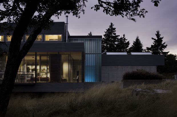 Twilight exterior view of a contemporary multi-level residence set among mature trees and ornamental grasses. The house features dark horizontal cladding, expansive glazed living areas with horizontal sun-shading louvers, a blue-grey vertical metal-clad volume, and a low flat-roofed concrete wing. Warm interior lighting glows through the windows.