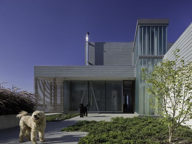 Exterior view of a contemporary low-rise residence with a layered flat-roof composition, dark horizontal cladding, extensive glazing, vertical glazed screening, and horizontal louvres. A landscaped entry garden and paved path lead toward the main entrance. The image is taken outdoors and primarily focuses on the building facade and approach.