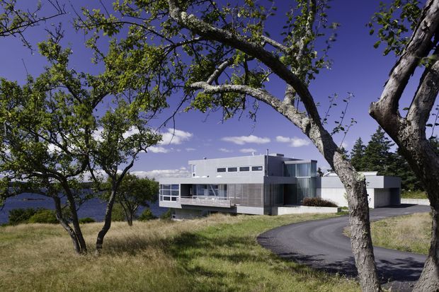 A modern, low-profile multi-level house set in a landscaped, gently sloping site. The building features crisp white rendered volumes, dark horizontal cladding, extensive glazing, and a flat roofline. A curved asphalt driveway approaches the house, while mature trees and dry grass frame the view; a body of water is visible in the distant background.