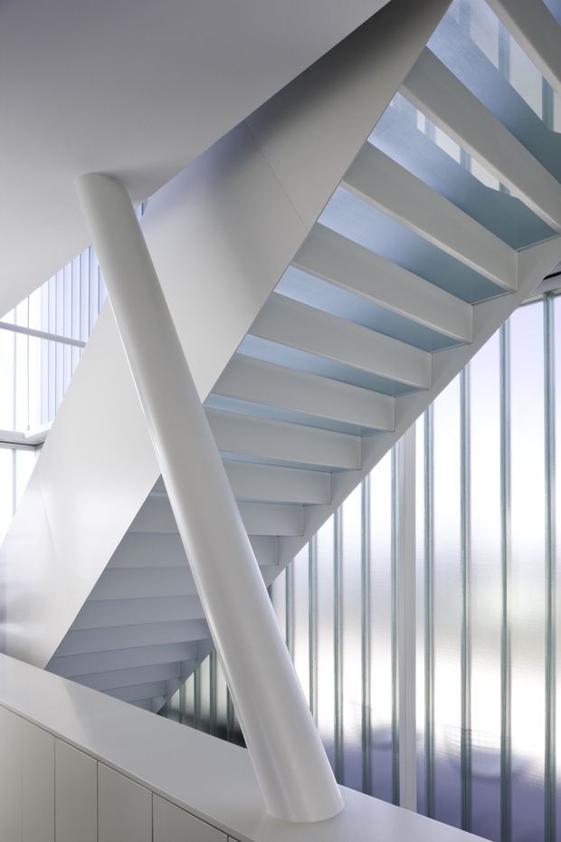 A bright, minimalist stairway with white sculptural stringer and diagonal support, floating white steps, and tall translucent vertical glazing that floods the stairwell with cool natural light. The composition emphasizes clean geometric lines, open structure, and a contemporary architectural interior.