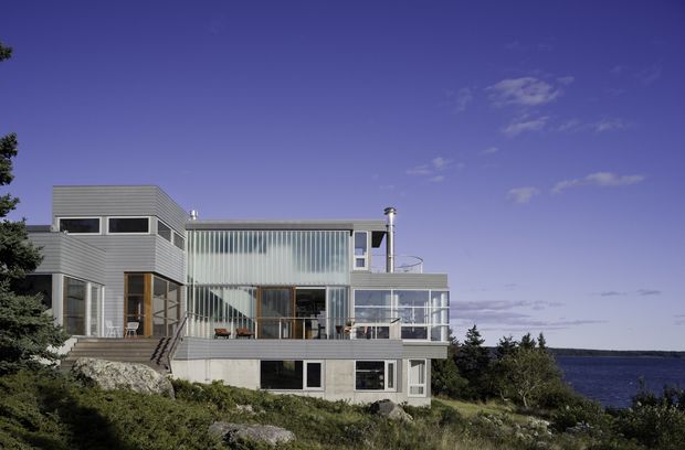 A modern multi-level coastal house set on a rocky, vegetated site beside the sea. The building has a highly articulated composition with horizontal grey cladding, extensive floor-to-ceiling glazing, translucent vertical screening, balconies, terraces, and a rooftop cylindrical chimney or vent element. The image is photographed from outside, with the residence and waterfront setting dominating the frame.