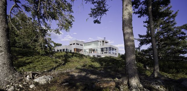 A modern multi-level building is partially visible through a wooded, rocky landscape. The structure features crisp white and grey volumes, extensive floor-to-ceiling glazing, projecting balconies or terraces, and a flat roofline. Tall mature trees frame the view, with exposed rock and natural ground in the foreground.