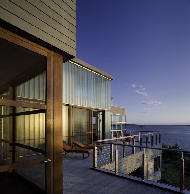 Exterior view of a contemporary coastal house at dusk, showing a timber-clad and glazed facade opening onto a spacious timber deck with a slim metal-and-glass balustrade. The building projects toward a calm body of water, with a long narrow pier visible beyond. Strong horizontal roof and cladding lines, extensive glazing, and warm interior light create a refined modern architectural composition.