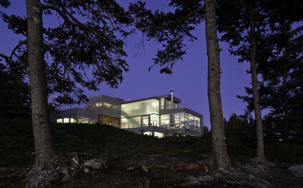 Dusk exterior view of a large contemporary multi-level house nestled among mature trees on a wooded, sloping site. The building features extensive floor-to-ceiling glazing, illuminated interiors, projecting volumes, flat rooflines, and multiple terraces or balconies. Dark tree trunks and rocky ground frame the warmly lit modern residence against a deep blue-purple sky.