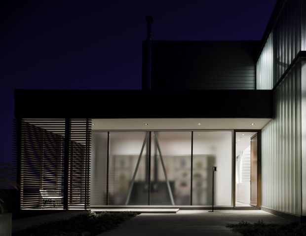 Night-time view of a modern, flat-roofed building entrance and courtyard. The facade combines extensive floor-to-ceiling glazing, dark horizontal cladding, vertical translucent ribbed panels, and a screened section of horizontal timber or metal battens. Interior furnishings are softly visible through the glazed wall, while recessed soffit lighting and subtle pathway lighting illuminate the entry.