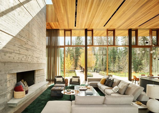 A spacious, light-filled contemporary living room with a large concrete fireplace wall, extensive floor-to-ceiling timber-framed glazing, and a warm timber-lined ceiling. A large sectional sofa, lounge chairs, coffee tables, and a deep green rug face the fireplace and wooded garden beyond. The room combines natural materials, strong horizontal and vertical lines, and an indoor-outdoor connection.