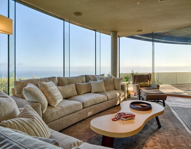 A spacious, refined living room with a large neutral sectional sofa, sculptural organic-shaped coffee table, armchairs, and a broad rug. Full-height glazing wraps around the room, opening to expansive ocean views and a covered terrace. A concrete column and smooth concrete ceiling reinforce the contemporary architectural setting.