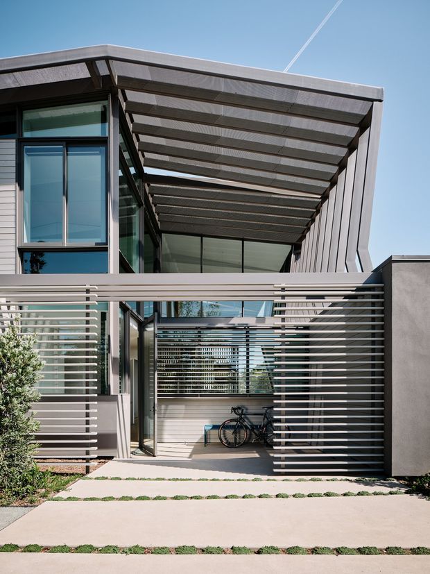 Exterior view of a contemporary multi-level residence with extensive glazing, horizontal metal privacy screens, a deep timber-toned pergola or covered walkway, and a concrete-paved entrance path. A bicycle is stored beneath the screened entry structure, with planting at the edges.