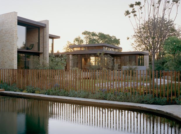 A contemporary low-rise residential property viewed from across a reflective pond. The building combines pale masonry volumes, dark framed floor-to-ceiling glazing, timber-toned screens, and broad low-pitched roof forms. A vertical timber fence and landscaped planting separate the house from the water, creating a calm, natural setting.