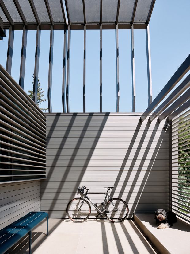 A bright, modern semi-enclosed outdoor courtyard or terrace framed by vertical and horizontal timber or metal screening. A slatted overhead pergola casts strong linear shadows across grey horizontal-clad walls and pale paving. The space includes a bicycle, a small dog, a blue bench or outdoor seat, and layered privacy screens, with clear sky visible above.