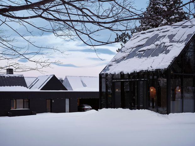 Snow-covered modern residential buildings in a winter landscape. The foreground structure is a dark-framed glazed pavilion or extension with a steep pitched roof, while adjacent dark-clad buildings with pitched roofs sit behind it. Heavy snow covers the roofs and ground, with warm interior lighting visible through the glazing.