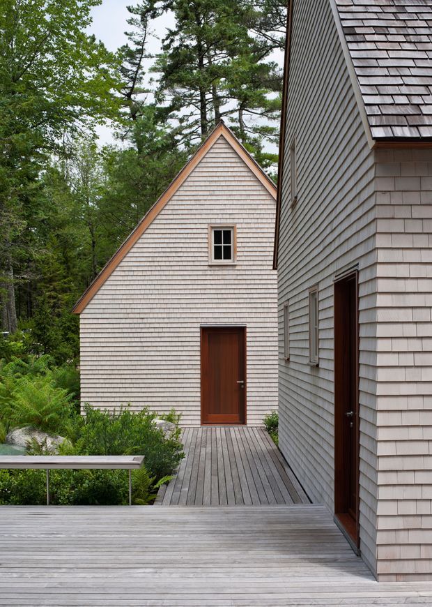 Exterior architectural view of a cluster of shingle-clad buildings connected by a broad timber deck and walkway. The buildings have steep gabled roofs, pale grey timber shingles, simple dark-trimmed windows, and dark timber doors. Dense woodland surrounds the deck, creating a secluded natural setting.