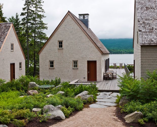 Exterior view of a group of shingle-clad buildings arranged around a landscaped gravel and stone path. The central building has a steep gabled roof, small-paned windows, a dark timber door, and a chimney, with timber decking leading toward a partially visible waterfront. Naturalistic planting, boulders, and mature trees frame the scene, with forested hills across the water.