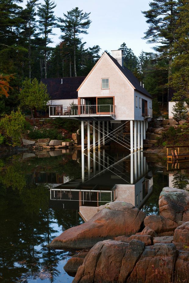 Exterior view of a compact, elevated house set among mature trees and large exposed rock formations beside a calm pond. The steep gable-fronted structure has pale shingle cladding, dark roofing, a chimney, and a projecting deck with simple metal railings. The building is supported above the water on slender columns with diagonal bracing, and its reflection is clearly visible in the pond.