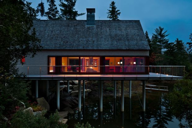 A modern elevated house at dusk, surrounded by dense evergreen trees and positioned over a reflective body of water or rocky natural terrain. The building has a broad steeply pitched shingle roof, dark horizontal cladding, extensive warm-lit glazing, and a long projecting deck supported by slender posts. The dramatic contrast between the cool blue exterior light and warm interior illumination gives the scene a serene yet atmospheric character.