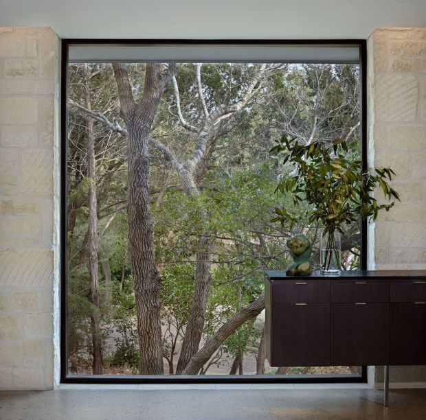 Interior view focused on a large picture window set into pale textured stone walls, framing dense natural woodland outside. A dark timber sideboard with slim metal legs and a leafy indoor plant occupies the lower right, creating a strong contrast with the light stone and muted forest view.