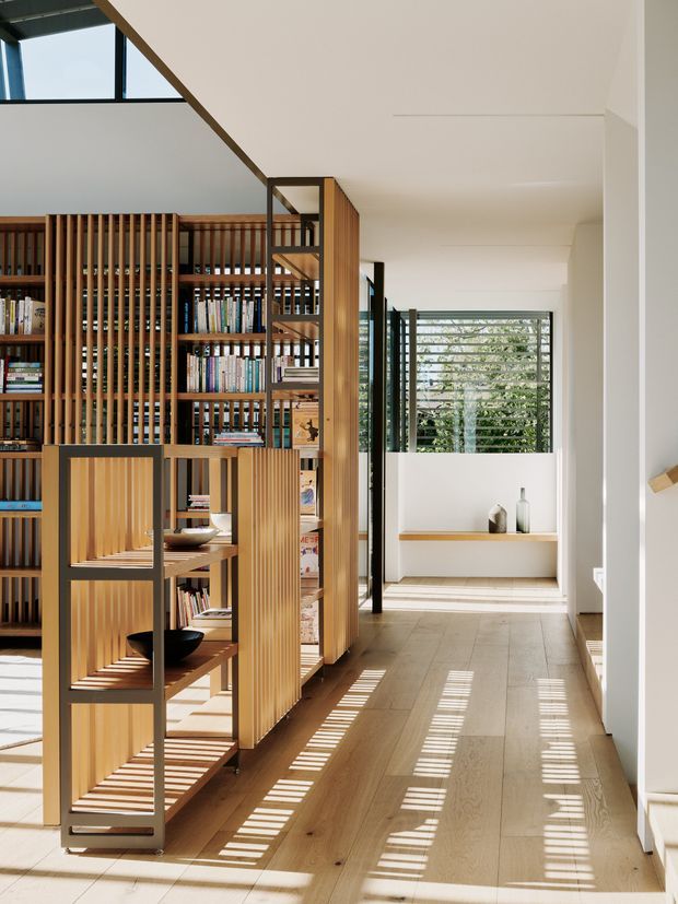 A bright contemporary interior hallway or living area featuring extensive custom timber shelving and vertical timber screening, filled with books and display objects. A freestanding timber-and-dark-metal shelving unit sits in the foreground, while a long timber floor leads toward a glazed opening with horizontal external screening. Strong sunlight creates linear shadow patterns across the floor, and the space has a light, open, carefully detailed feel.