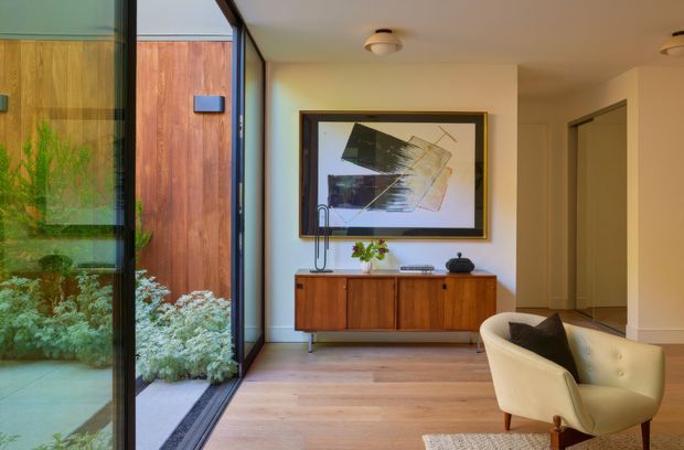 A refined mid-century-inspired living room or entry space with warm timber flooring and a matching low wooden sideboard, cream upholstered lounge chair, abstract framed artwork, and simple ceiling lights. Full-height glazing on the left looks onto a planted courtyard and a timber-clad exterior wall, while a hallway or adjoining room is visible at right. The interior has a calm, warm, understated character with strong indoor-outdoor connection.