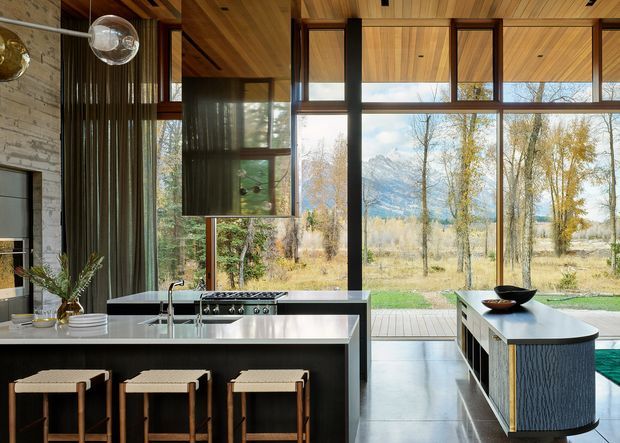 A sophisticated contemporary kitchen with a large central island and three woven-seat stools, a secondary cooking bench, dark cabinetry, pale stone benchtops, and extensive floor-to-ceiling glazing. Warm timber ceilings and window framing contrast with dark vertical elements, while a mountain and woodland landscape is visible beyond the glazed facade.