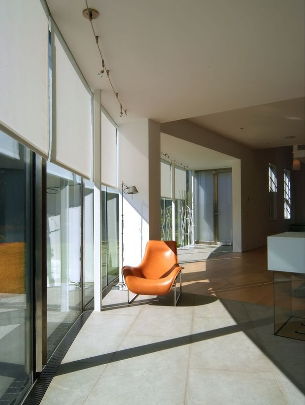 A bright, minimalist interior with expansive floor-to-ceiling glazing along one side, translucent roller blinds, pale large-format flooring, and a warm neutral ceiling and wall palette. Strong sunlight creates geometric shadows across the floor. An orange sculptural lounge chair sits beside the glazing, while a white built-in island or cabinet is visible at right. Track lighting and a slim floor lamp add understated illumination.