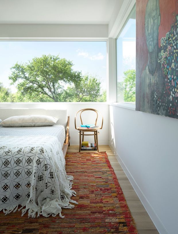Bright, compact bedroom with a single bed dressed in a white patterned fringed coverlet, a simple wooden bentwood chair, and a colourful woven runner rug. Large corner windows provide abundant daylight and views of leafy trees. White walls and trim create an airy backdrop, while a large colourful artwork adds a bold feature to the right wall.