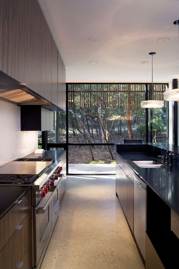 A contemporary galley kitchen with dark vertical-grain timber cabinetry, stainless-steel appliances, black benchtops, and a large glazed opening at the rear. Full-height windows and vertical timber screening connect the kitchen to a wooded outdoor setting. The space has a clean linear layout, recessed ceiling lights, pendant lighting, and a polished functional finish.
