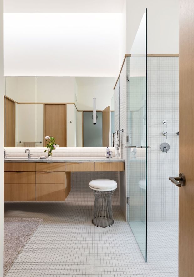 Bright contemporary bathroom with a floating timber vanity, integrated basin, large mirror, small-grid light tile flooring and shower walls, frameless glass shower screen, chrome fixtures, and a sculptural wire-frame stool. A timber door and warm wood trim add contrast to the predominantly white space.