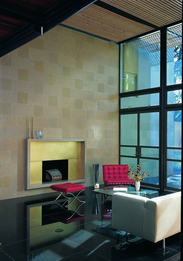 A sophisticated double-height living room with a monumental tiled stone fireplace, glossy black floor, exposed timber slat ceiling, and tall black-framed glazing. The room is furnished with a sculptural white armchair, vivid red tufted lounge chairs and ottoman, and a transparent glass coffee table. Strong daylight enters through the expansive windows, creating reflections across the polished floor.