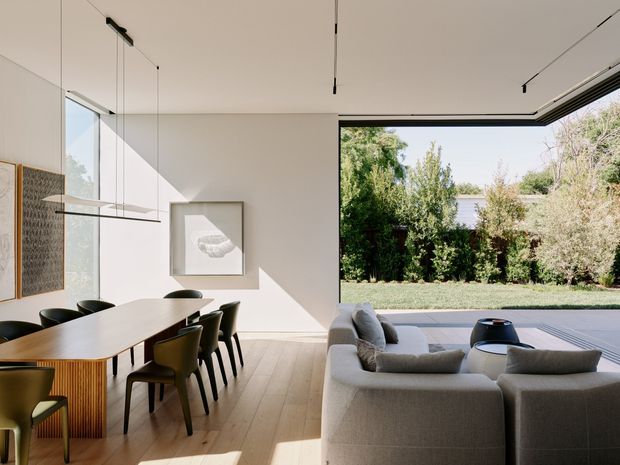 A bright, open-plan dining and living area with a long timber dining table and dark upholstered chairs in the foreground, and a large light-grey sectional sofa facing broad floor-to-ceiling glazing. The room has pale timber flooring, white walls and ceiling, minimalist linear pendant and track lighting, framed wall art, and strong indoor-outdoor connection to a landscaped garden. The architecture is clean-lined and contemporary with expansive glazing and a high, airy ceiling.