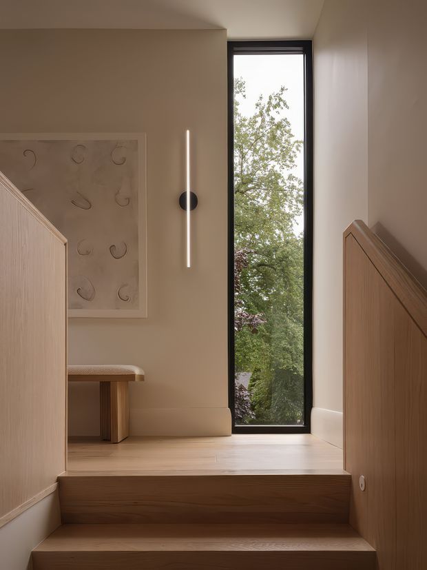 A warm, minimalist stair landing with pale timber steps and timber balustrades, beige painted walls, a tall narrow black-framed window, and a slim vertical wall light. A small timber bench and abstract artwork add subtle detail, while leafy trees are visible outside.