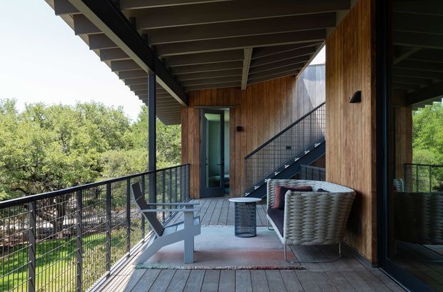 A covered elevated outdoor terrace or balcony with timber decking, vertical timber-clad walls, dark steel framing and railings, and a visible exterior stair. Woven outdoor lounge chairs and a small side table sit on a rug beside large dark-framed glazing. Dense leafy vegetation is visible beyond the balustrade.