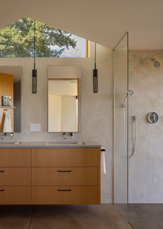 A contemporary bathroom with a floating warm-wood vanity, grey stone-look benchtop, twin basins and mirrors, and a large frameless glass walk-in shower. The room has plastered or microcement-style walls, dark flooring, black pendant lights, and a high horizontal window looking onto trees.