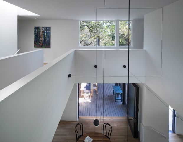 Interior view of a bright, minimalist multi-level home focused on a stairway and open void. White walls and balustrades frame a lower dining area with a timber table and black chairs, while tall black pendant lights hang through the void. Large windows overlook trees, and an outdoor timber deck is visible beyond the lower glazing.
