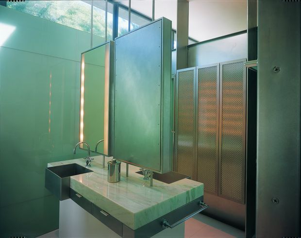 A contemporary communal bathroom with a long pale stone double vanity, integrated rectangular basins, exposed chrome taps, and under-counter drawers. Large frameless mirrors and translucent green-tinted partitions surround the sinks, while perforated metal privacy screens define the stalls in the background. High windows bring in soft natural light, creating a cool, slightly industrial atmosphere.